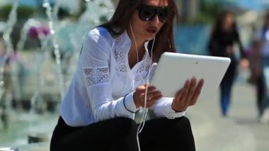 İş kadını fıskiyelerin yanında oturur, kahve içer ve tablet bilgisayarla iletişim kurar. Beyaz tablet bilgisayarlı gözlüklü çekici bir kadın Skype üzerinden iletişim kuruyor.