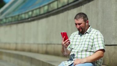 Komik adam akıllı telefonla iletişim kuruyor. Akıllı telefonlu neşeli adam Skype üzerinden iletişim kuruyor ve el kol hareketi yapıyor. Elinde kırmızı cep telefonu olan, kareli tişörtlü, şişman, şen şakrak adam.