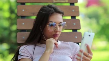Genç güzel kız parkta bankta oturur ve müzik dinler. Büyük gözlüklü çekici bir kadın elinde kulaklıklarla telefon tutuyor ve müzik dinliyor.