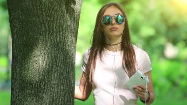 Genç güzel kız ağacın yanında duruyor, elinde akıllı bir telefon tutuyor, müzik dinliyor ve dans ediyor. Güneş gözlüklü çekici bir kadın doğada müzik dinliyor, arka planda titreşen güzel bir güneş.