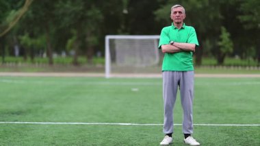 Futbol antrenörü futbol sahasında, beden eğitimi öğretmeni. Yeşil tişörtlü adam futbol sahasında duruyor ve kameraya bakıyor.