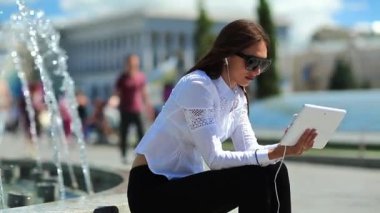 İş kadını çeşmelerin yanında oturur ve tablet bilgisayar kullanır. Elinde beyaz tablet bilgisayarla gözlüklü çekici bir kadın.