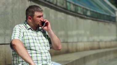 Adam akıllı telefondan konuşuyor. İşadamı basamaklarda oturur ve akıllı telefon aracılığıyla iletişim kurar. Adam cep telefonuyla konuşuyor ve el kol hareketi yapıyor. Elinde kırmızı cep telefonu olan şişman bir adam.