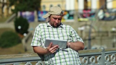 Beyzbol şapkalı şişman adam tablet bilgisayar kullanıyor. Kareli gömlekli çocuk şehirdeki tablet bilgisayarıyla çalışıyor.