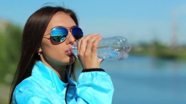 Güneş gözlüklü çekici kız şişeden temiz maden suyu içiyor. Mavi spor ceketli güzel kadın su içer ve etrafa bakar.