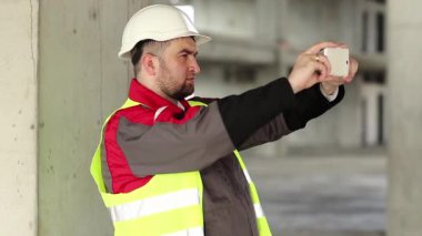 İnşaat alanındaki inşaatçı akıllı telefondaki videoları kaydediyor. Proje alanında akıllı telefonu olan beyaz şapkalı bir işçi