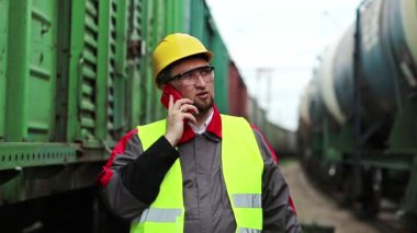 Demiryolu işçisi yük trenlerinin yanında cep telefonuyla konuşuyor. Demiryolu çalışanı demiryolu hattında çalışıyor. Cep telefonuyla konuşuyor. İşyerinin müdürü, nakliye istasyonundaki akıllı telefon aracılığıyla iletişim kuruyor.