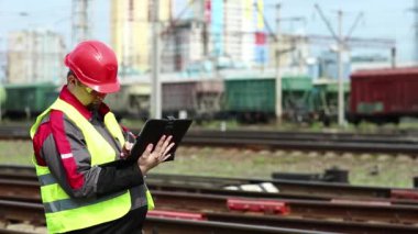 Yük terminalinde tablet bilgisayarlı demiryolu işçisi. Demiryolu çalışanı tabletine not alıyor. Üniformalı ve kırmızı şapkalı demiryolu işçisi tablet bilgisayarla çalışıyor.