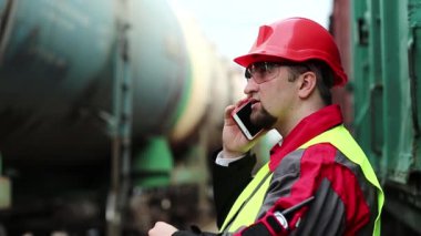 Demiryolu işçisi yük trenlerinin yanında cep telefonuyla konuşuyor. Demiryolu çalışanı demiryolu hattında çalışıyor. Cep telefonuyla konuşuyor. İşyerinin müdürü, nakliye istasyonundaki akıllı telefon aracılığıyla iletişim kuruyor.