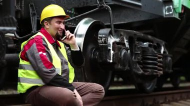 Neşeli demiryolu işçisi cep telefonuyla konuşuyor. Demiryolu çalışanı iş kıyafetleri içinde yük vagonunun yanında oturur ve cep telefonuyla konuşur.