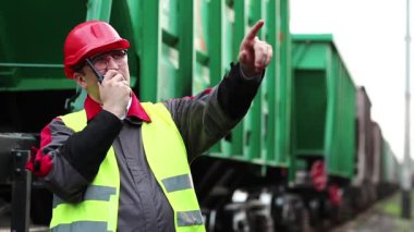 Trenlerin yanında taşınabilir radyo vericisi olan bir demiryolu işçisi. Demiryolu çalışanı, nakliye istasyonunun yanındaki demiryolu hattında portatif radyo vericisi aracılığıyla konuşuyor. İşçi radyo istasyonu aracılığıyla iletişim kuruyor.