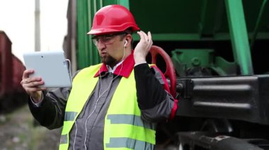 Demiryolu çalışanı, malzeme treninin yanındaki tablet bilgisayarla iletişim kuruyor. Yük istasyonundaki demiryolu trafiğinin müfettişi tablet bilgisayarla konuşuyor
