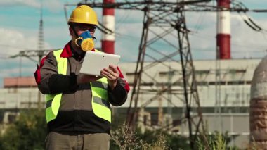 Elektrik santralindeki tablet bilgisayarlı gaz maskeli adam. Güç mühendisliği uzmanı solunum cihazında ve sarı kasketli ve kişisel bilgisayarlı ısı santralinde çalışıyor.
