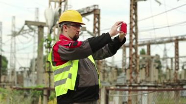 Elektrikçi akıllı telefondan video kaydediyor. Elektrik santralinde çalışan, sarı şapkalı, akıllı telefonlu bir işçi.