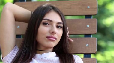 Şehir parkındaki bankta oturan güzel kadın kameraya bakıyor ve gülümsüyor. Uzun saçlı çekici kız halka açık bir bahçede bankta oturuyor.