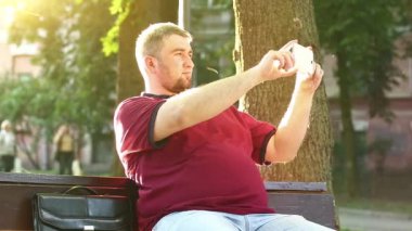 İş adamı bankta oturur ve akıllı telefondaki videoları kaydeder. Adam cep telefonuyla fotoğraf çekiyor.