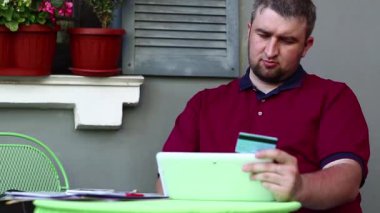 Adam yaz restoranında masada oturuyor, elinde beyaz tablet bilgisayar ve mavi kredi kartı tutuyor, veri giriyor. İşadamı kredi kartıyla online alışveriş yapıyor, kredi kartıyla alışveriş yapıyor.