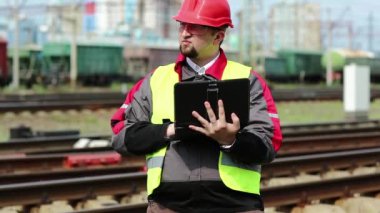 Tren terminalinde tablet bilgisayarlı demiryolu işçisi. Üniformalı ve kırmızı şapkalı demiryolu işçisi tablet bilgisayarla çalışıyor. Demiryolu çalışanı tabletine notlar alıyor.