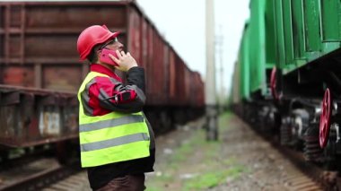 Demiryolu işçisi, yük trenleri arasında cep telefonuyla konuşur. Demiryolu çalışanı demiryolu hattında çalışırken cep telefonundan konuşuyor. Demiryolu işçisi, nakliye istasyonunda akıllı telefondan iletişim kuruyor.