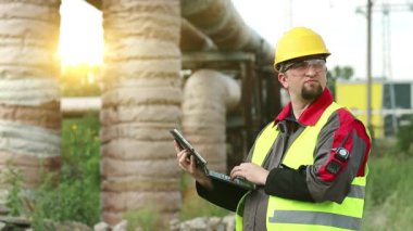 Ana boru hattının yakınındaki ısı elektrik santralinde tablet bilgisayarı olan bir işçi. Sarı başlık mühendisliği uzmanı tabletle çalışıyor, arka planda güzel bir güneş var.