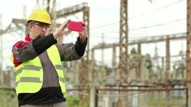 İşçi akıllı telefondan video kaydediyor. Elektrik santralinde akıllı telefonlu sarı şapkalı güçlü adam.