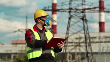 Elektrik santralindeki gaz maskeli adam iş belgelerine not alıyor. Elektrik mühendisliği uzmanı. Solunum cihazında ve sarı kaskette. Belgelerde yazıyor.