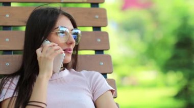 Şehir parkındaki bankta oturan ve cep telefonuyla konuşan güzel bir kadın. Çekici kız cep telefonuyla konuşuyor. Beyaz akıllı telefonlu bir kadın. Akıllı telefonlu iş kadını halka açık bir bahçede oturuyor.