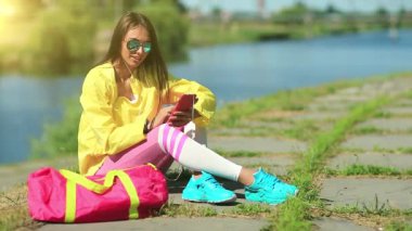 Sporcu kadın göl kenarında oturuyor ve akıllı telefonunu kullanıyor. Sarı spor ceketli, pembe spor çantalı güneş gözlüklü güzel bir kız göletin yanında oturuyor ve cep telefonu kullanıyor. Cep telefonlu spor elbiseli bir kadın.