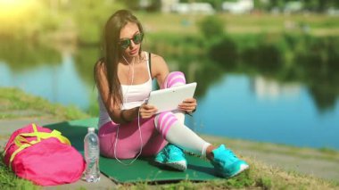 Sporcu kadın gölün yanındaki yeşil mindere oturur ve tablet bilgisayarla iletişim kurar. Güzel kız gölün yanında oturuyor ve tablet bilgisayarla konuşuyor. Spor kıyafetli bir kadın tablet bilgisayarla Skype üzerinden iletişim kuruyor.