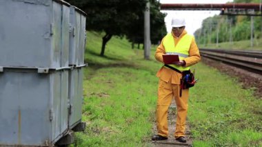 Elektrikçi belgelerine not alıyor. Demiryolu çalışanı sarı üniformalı demiryolu hattında. Sarı üniformalı demiryolu işçisi ve elinde belgelerle beyaz şapkalı.