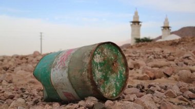 Demir fıçı taşlı zeminde, arka planda iki minaresi olan bir camide yatıyor. Cami ve petrol varili. Ürdün, Akabe 'deki dağda eski, paslı bir fıçı. Boş fıçı, yakıt yok.