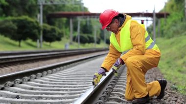 Demiryolu işçisi sarı üniformalı ve demiryolu hattında ölçüm aleti olan. Demiryolu işçisi kırmızı şapkalı tren raylarında oturur ve trene bakar. Seviye ölçüm aletine sahip işçiler