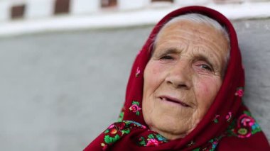 Kıdemli kadın kameraya bakar ve gülümser. Kırmızı başörtülü yaşlı kadın evinin gri duvarının yanında oturuyor ve kameraya bakıyor. Kadın kameraya bakıyor ve gülümsüyor, buruşuk surat.