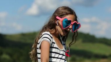 Akıllı telefonlu güzel kız çayırda duruyor, müzik dinliyor, dans ediyor ve şarkı söylüyor. Büyük gözlüklü, kalp şeklinde akıllı bir kız müzik dinler, şarkı söyler ve yeşil çimlerde dans eder.