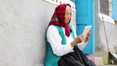 Yaşlı kadın bankta oturuyor ve tablet bilgisayarla iletişim kuruyor. Kıdemli kadın evinin yanındaki bankta oturuyor ve Skype 'ta tablet bilgisayarla konuşuyor. Tabletli Ukraynalı yaşlı kadın Skype üzerinden iletişim kuruyor