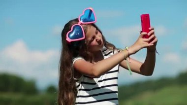 Uzun saçlı güzel bir kız akıllı telefonunda selfie çekiyor. Kalp şeklinde gözlüklü bir kız akıllı telefonunda selfie çekiyor. Kırmızı cep telefonlu güzel bir kız fotoğraf çekiyor. Kadın fotoğraf çeker.
