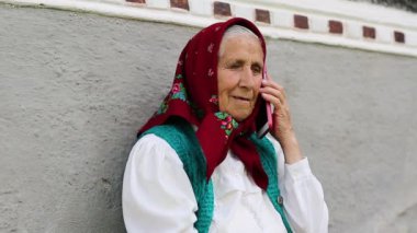 Yaşlı kadın evinin yanında oturuyor ve cep telefonuyla konuşuyor. Akıllı telefonlu bir kadın. Kırmızı akıllı telefonlu Ukraynalı yaşlı kadın.