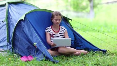 Kız turist çadırının yanındaki yeşil çimlerde oturuyor ve tablet bilgisayarına bakıyor. Tabletli kız, turist çadırının yanındaki yeşil çimlerin üzerinde oturuyor. Tablet pc ekranında kadın görünümü