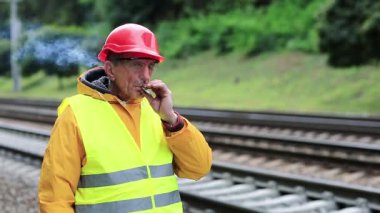 Demiryolu işçisi kırmızı başlıklı adam tren yolunda duruyor ve sigara içiyor. Demiryolunda sigara içen işçi. Sigara molası. Sarı üniformalı demiryolu işçisi demiryolu hattında duruyor.