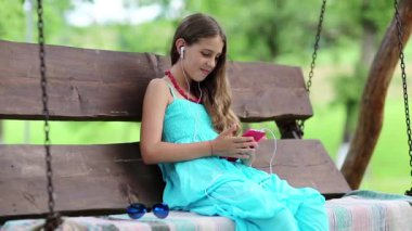 Mavi elbiseli güzel kız bahçede salıncakta oturuyor ve kırmızı akıllı telefondan konuşuyor. Kulaklığı ve akıllı telefonu olan çekici bir kız Skype üzerinden iletişim kuruyor. Akıllı telefonu olan bir kadın.