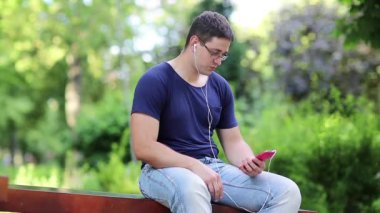 Adam kırmızı akıllı telefonla iletişim kuruyor. Genç adam şehir parkındaki bankta oturuyor ve akıllı telefondan konuşuyor. Adam halka açık bir bahçede bankta oturuyor ve cep telefonuyla Skype üzerinden iletişim kuruyor.