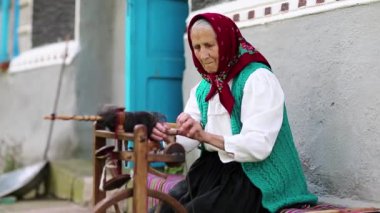 Yaşlı kadın bankta oturuyor ve dönen tekerlekle distaff ile çalışıyor. Ukraynalı yaşlı kadın evinin yanındaki bankta oturur ve iplik örer. Dönen tekerli bir kadın, el işi.