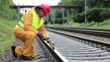 Demiryolu işçisi sarı üniformalı ve demiryolu hattında ölçüm aleti olan. Demiryolu işçisi kırmızı şapkalı tren raylarında oturur ve trene bakar. Seviye ölçüm aletine sahip işçiler