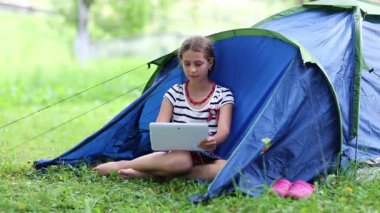 Güzel kız turist çadırının yanındaki çimlere oturur ve tablet bilgisayar kullanır. Tabletli kız, turist çadırının yanındaki yeşil çimlerin üzerinde oturuyor. Doğayla ilgili tabletli bir kadın.