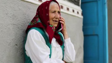 Yaşlı kadın evinin yanındaki bankta oturuyor ve cep telefonuyla konuşuyor. Kırmızı akıllı telefonlu Ukraynalı yaşlı kadın. Akıllı telefonu olan bir kadın.