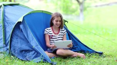 Güzel kız turist çadırının yanındaki çimlerde oturur ve tablet bilgisayarla iletişim kurar. Tabletli kız, turist çadırının yanındaki yeşil çimlerin üzerinde oturuyor. Tabletli güzel kız Skype üzerinden iletişim kuruyor.