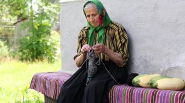 Ukraynalı yaşlı kadın örgü örüyor. Yaşlı kadın bankta oturup yün çoraplar örüyor. Yeşil başörtülü yaşlı kadın evinin yanında oturup çorap örüyor. Eşarplı kadın yün çorap örüyor.