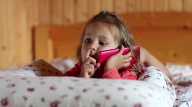 Kırmızı akıllı telefonlu komik küçük kız yatağa uzanıyor ve burnunu karıştırıyor. Küçük kız yastığa uzanıyor ve cep telefonunu tutuyor. Kırmızı cep telefonu olan küçük kız kanepede yatıyor.