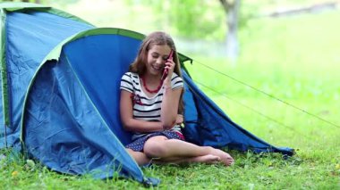 Kız turist çadırının yanındaki çimlerde oturur ve cep telefonuyla konuşur. Kırmızı akıllı telefonlu güzel kız mavi turist çadırının yakınındaki çimlerin üzerinde oturuyor. Güzel kız cep telefonuyla konuşuyor.
