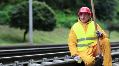Sarı üniformalı, elinde kürekle demiryolu işçisi demiryolu hattında oturuyor. Kırmızı şapkalı demiryolu işçisi raylara oturup trene bakıyor. Demiryolunda kürekli bir işçi. Demiryolu inşaatı